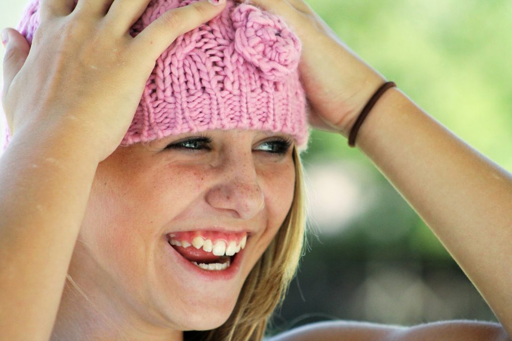 Dziewczynka w różowej czapce uśmiecha się i trzyma się rękami za głowę. Emocje radości.